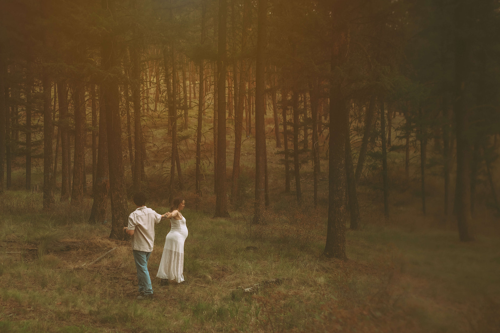 A mom to be leads her partner through the forest by the hand in a white maternity gown after exploring baby shower venues in Polson, MT