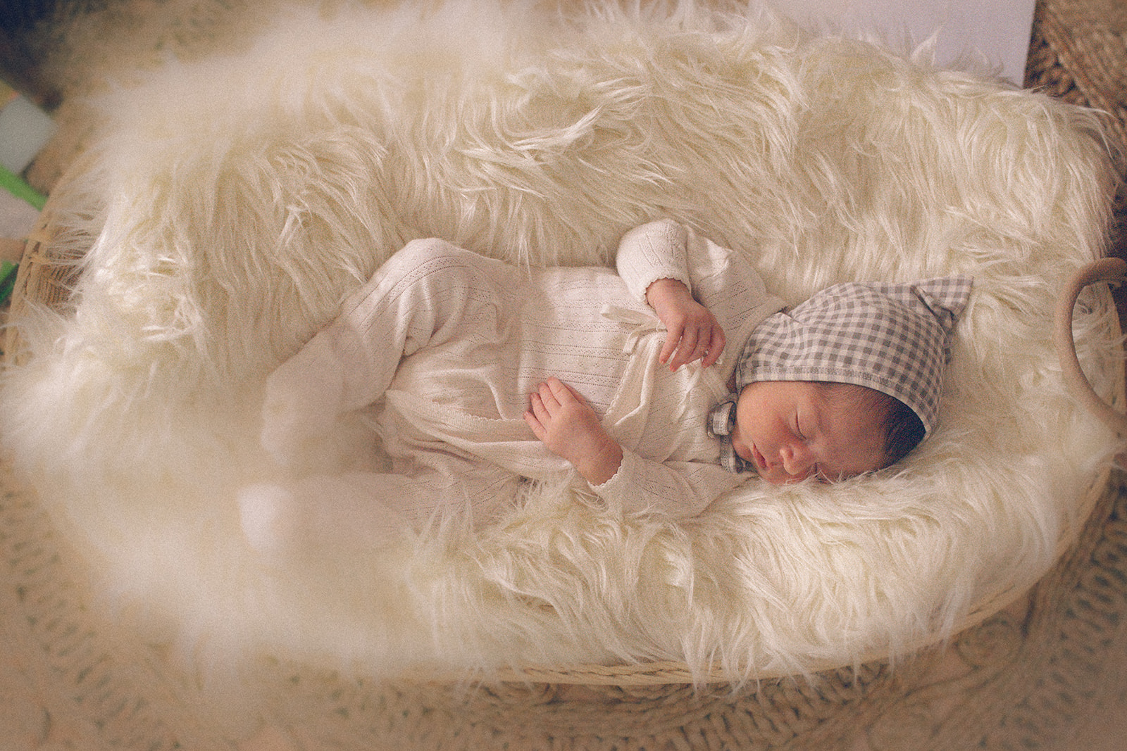 A look down at a newborn baby sleeping in a white onesie and sleep cap on a fur pillow in a basket