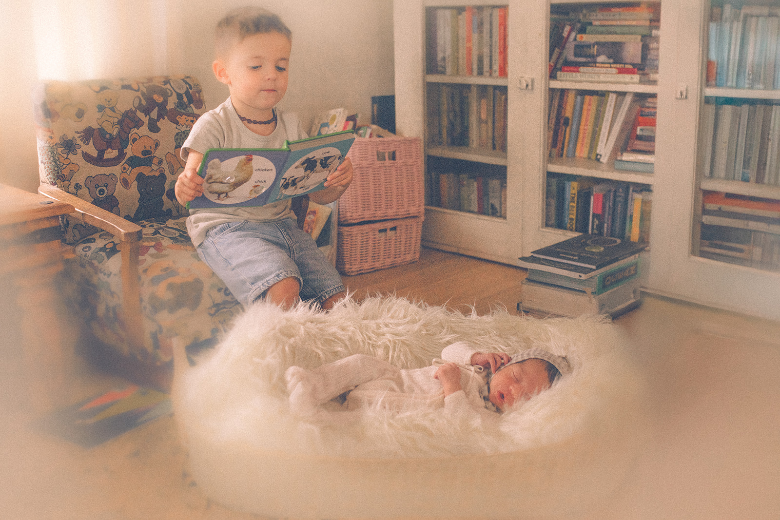 A toddler boy sits in a child's chair reading a book to his sleeping newborn baby sibling in front of him after visiting Briars Baby Boutique