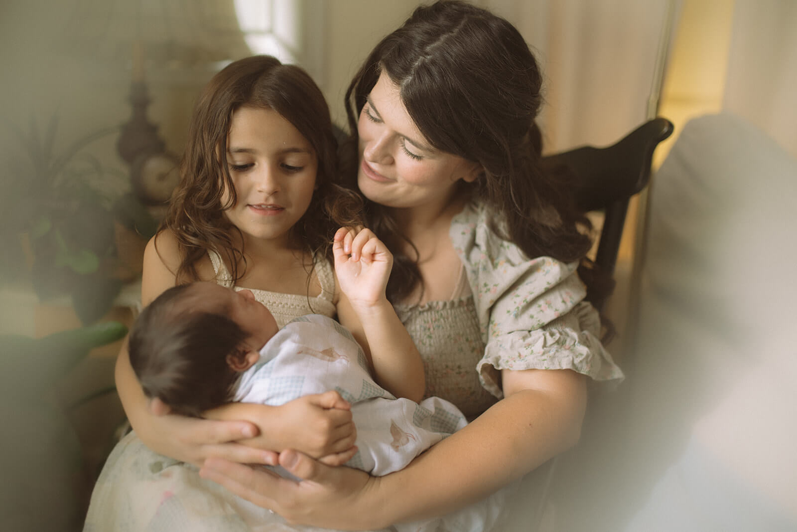 A happy mom in a flower print dress cradles her toddler daughter and newborn in her lap in a rocking chair after visiting handmade montana