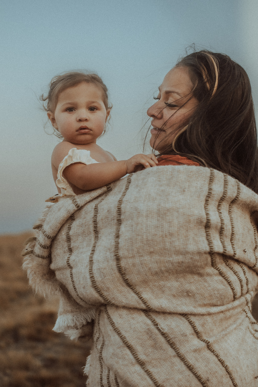 A happy mom wrapped in a tan blanket smiles at her toddler daughter in her arms outdoors