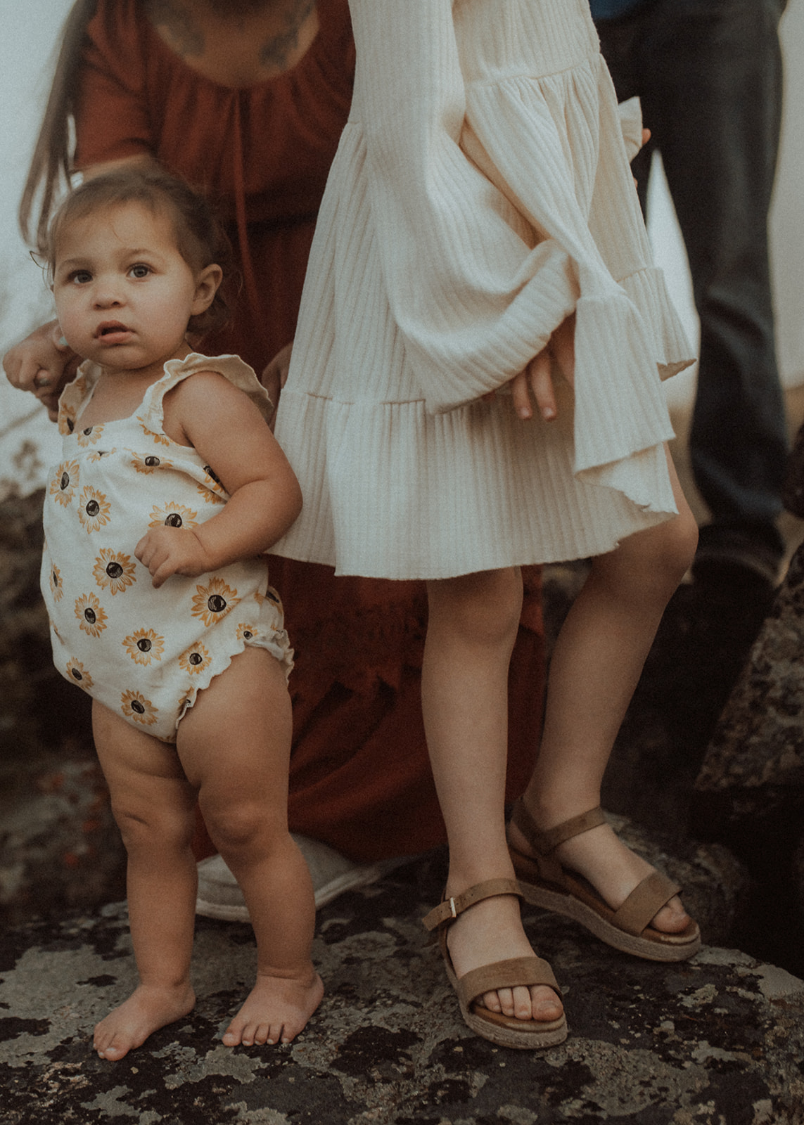 A toddler girl stands with big sister and mom and dad on a boulder in a yellow flower onesie after visiting head over heals in Polson