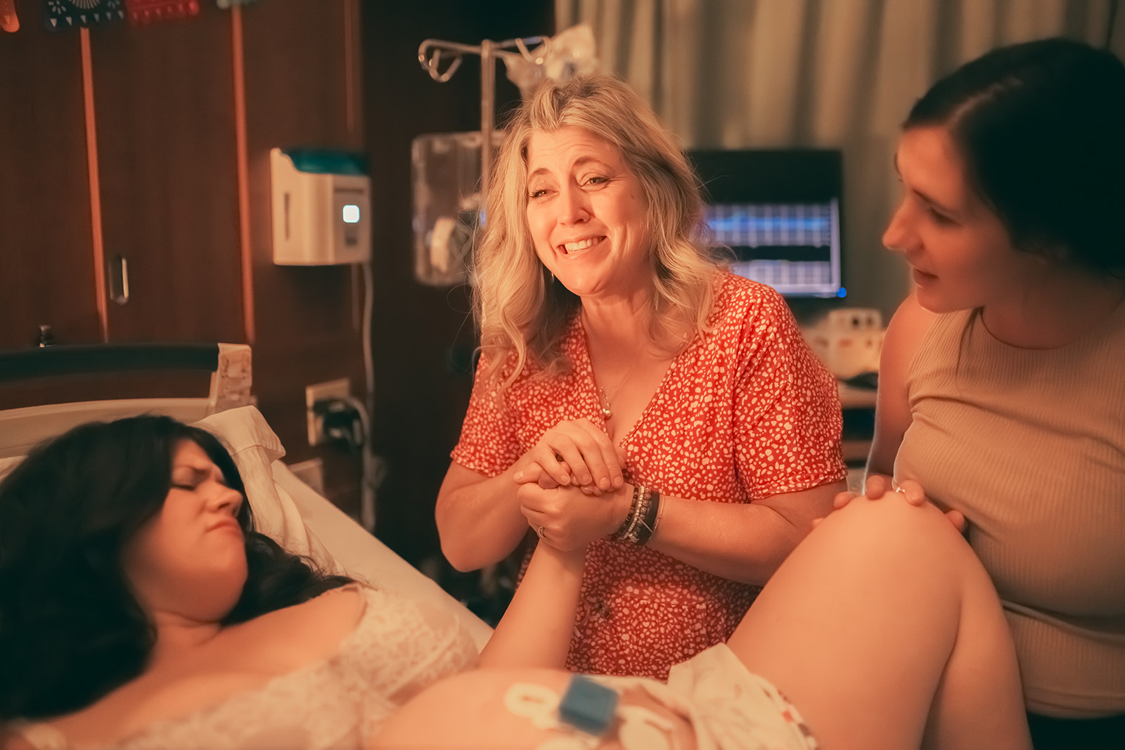A smiling woman in a red dress comforts a woman in labor in a hospital bed