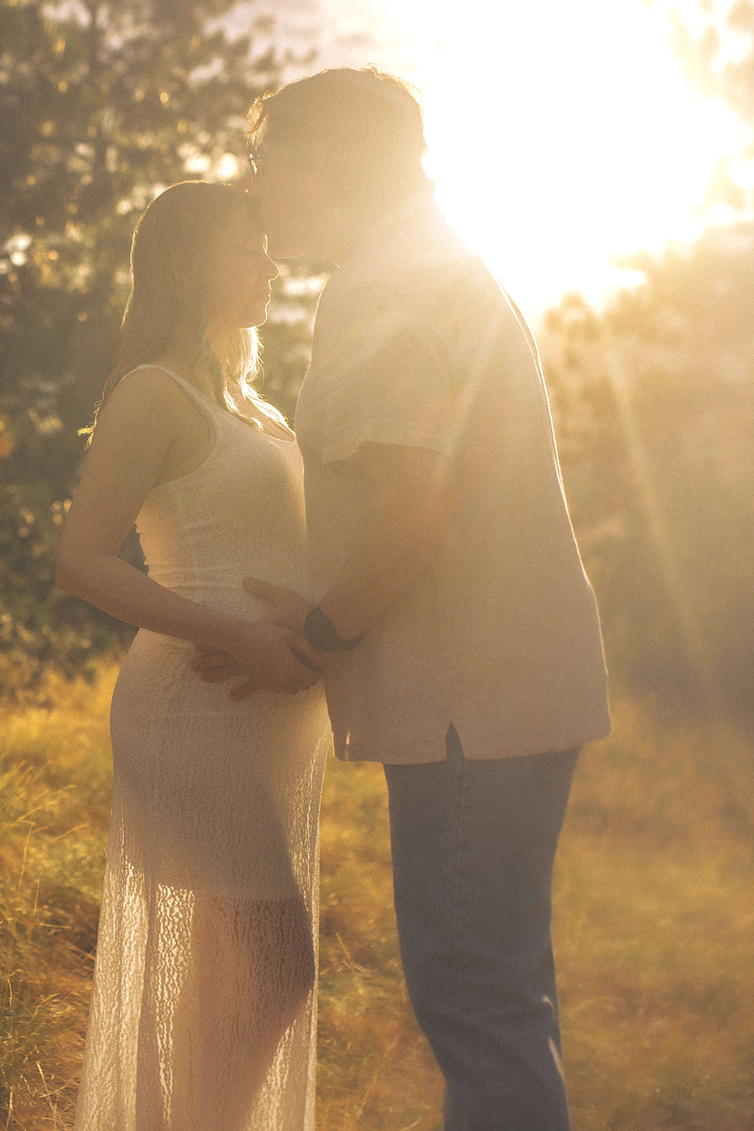 A mom kisses his expecting wife in a white maternity gown in the sun at sunset in a meadow
