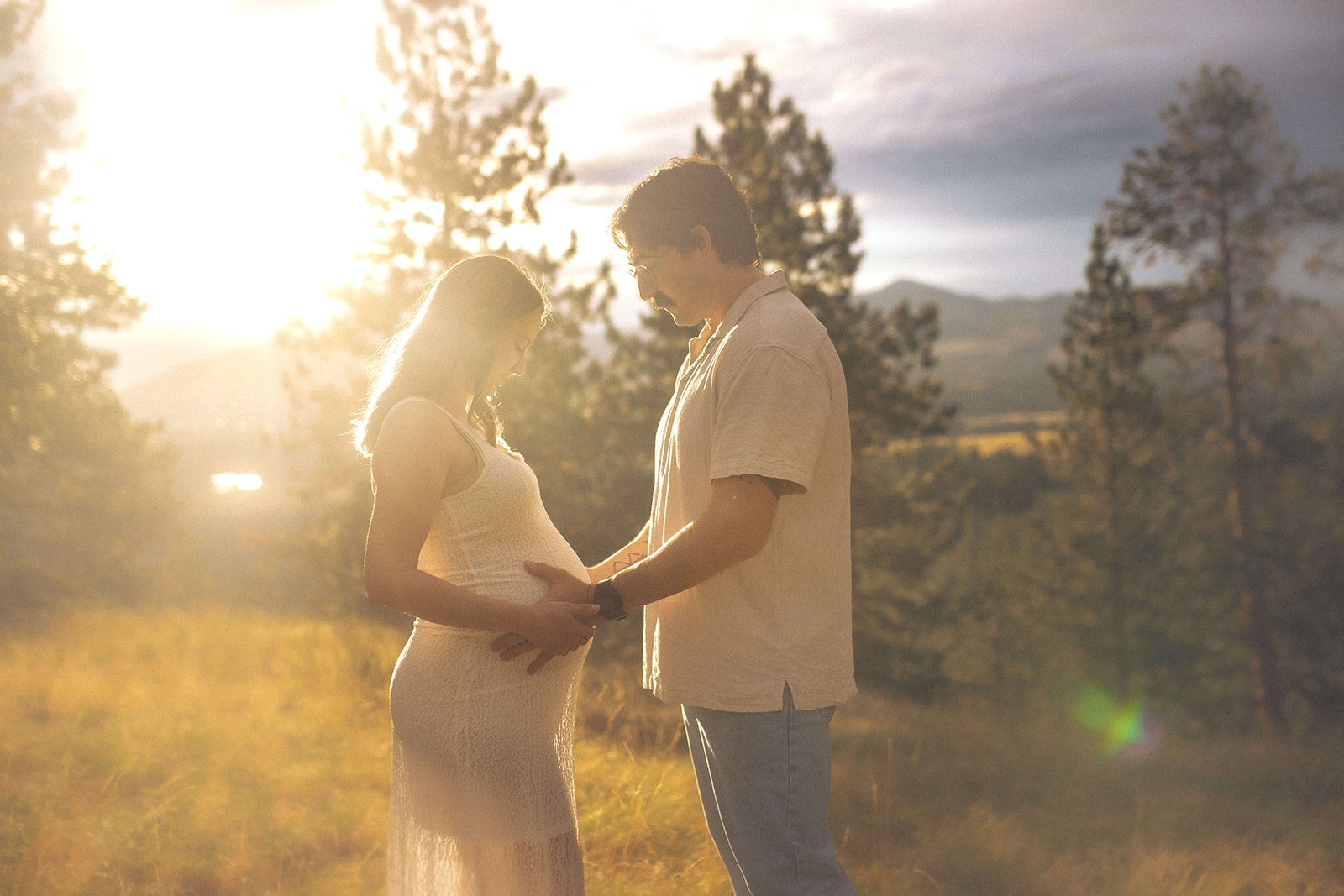 Smiling pregnant couple hold the bump while standing in a mountain meadow at sunset after a prenatal massage in Polson, MT