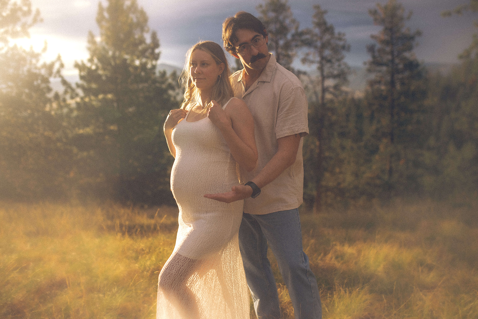 A man in a white shirt presents his pregnant wife in a white maternity gown in a meadow at sunset after a prenatal massage in Polson, MT