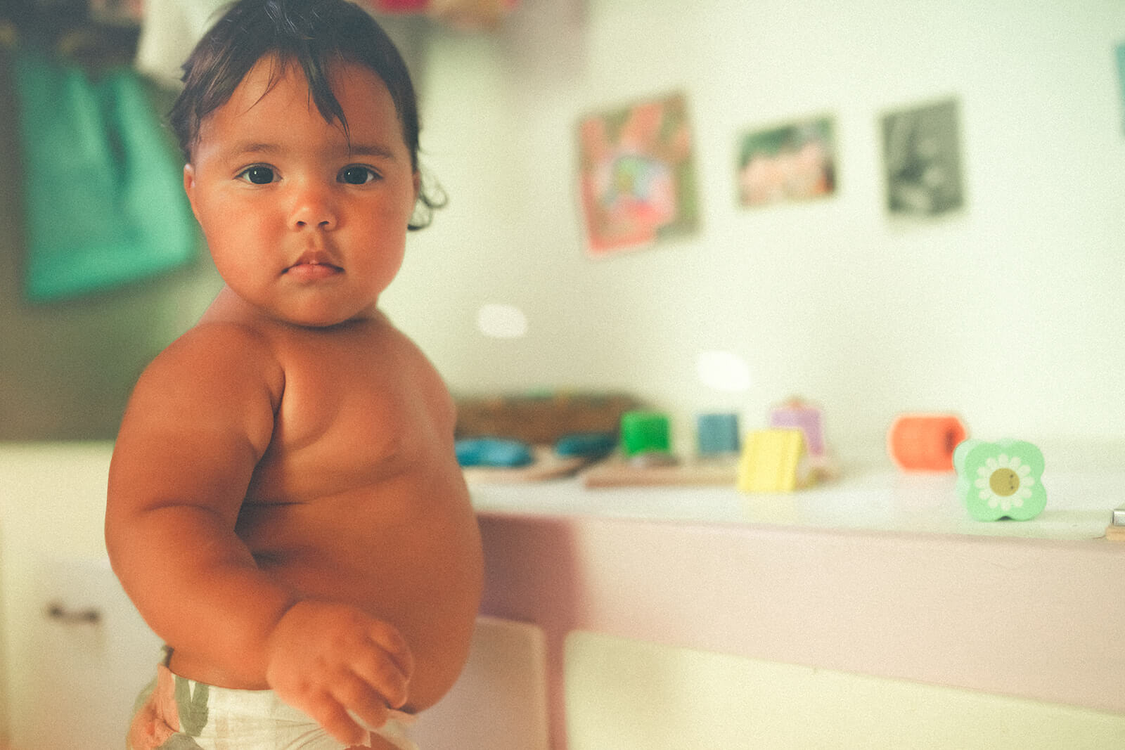 A baby in a diaper stands to play with small toys on a bench in a nursery