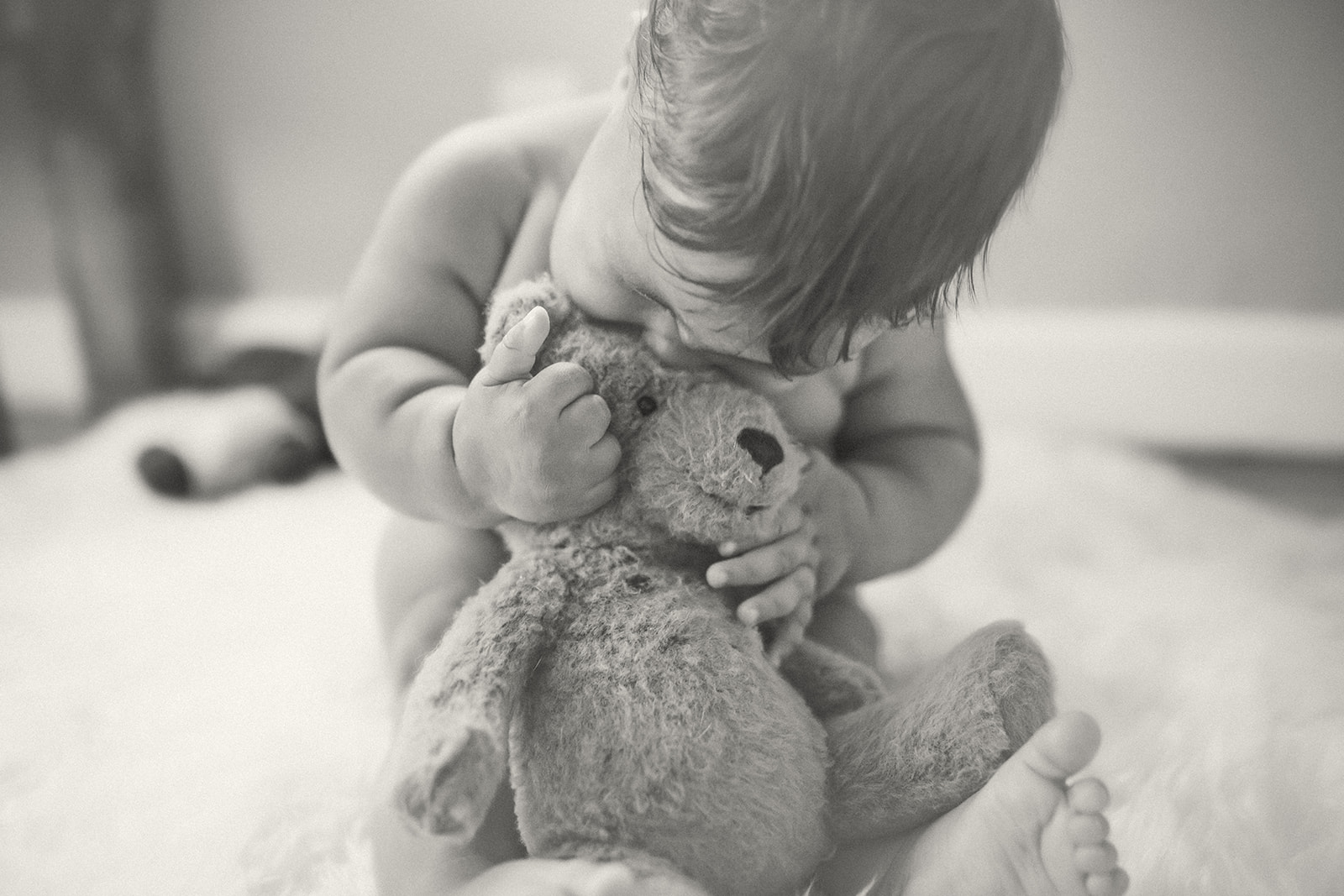 An infant snuggles a teddy bear in his lap while sitting on the floor in a living room in black and whtie