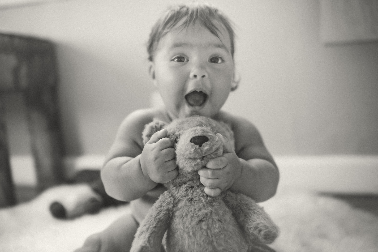 A silly baby makes faces while playing with a teddy bear on the living room floor in black and white after visiting daycares in Kalispell, MT