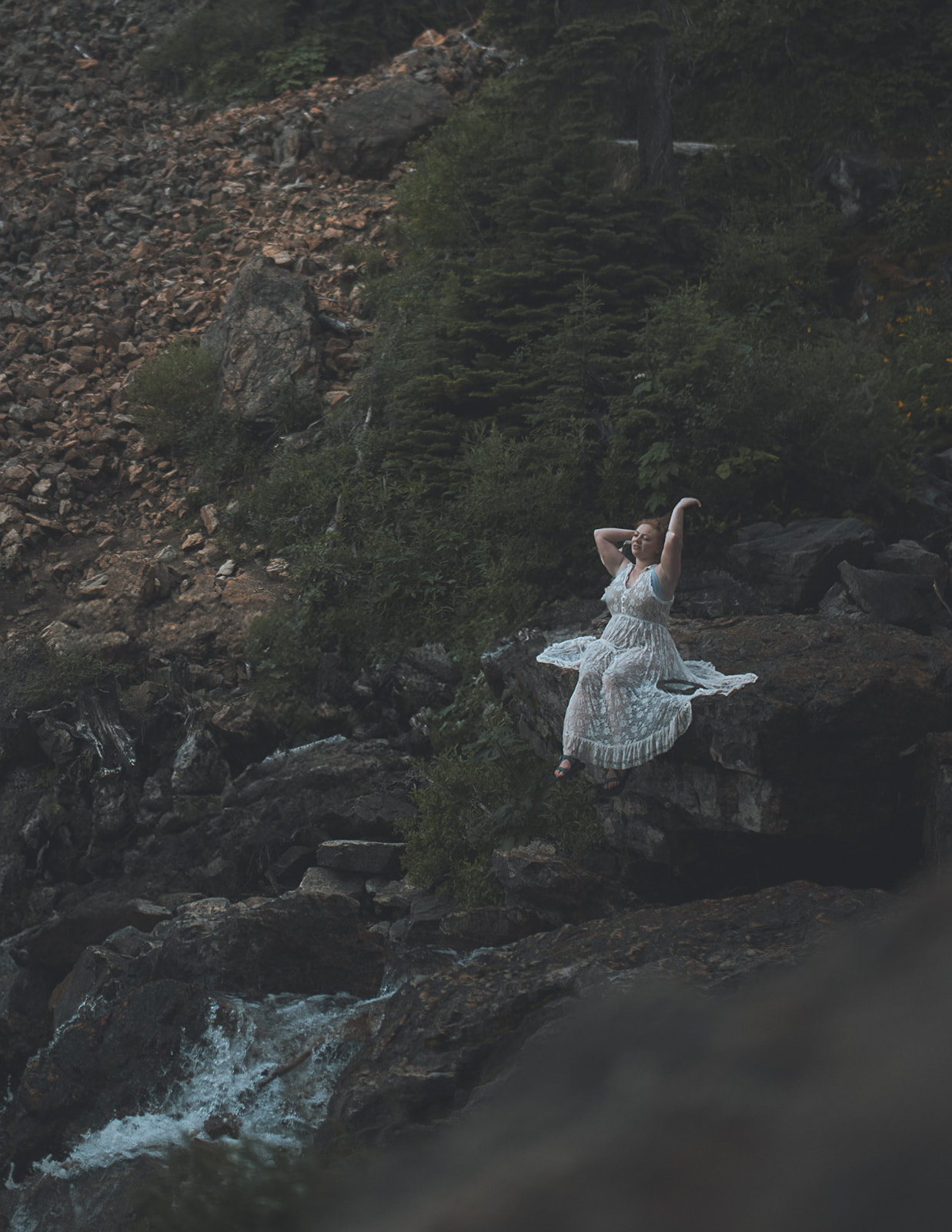 A woman adjusts her hair while sitting on a large boulder on a waterfall while wearing a white lace coverup