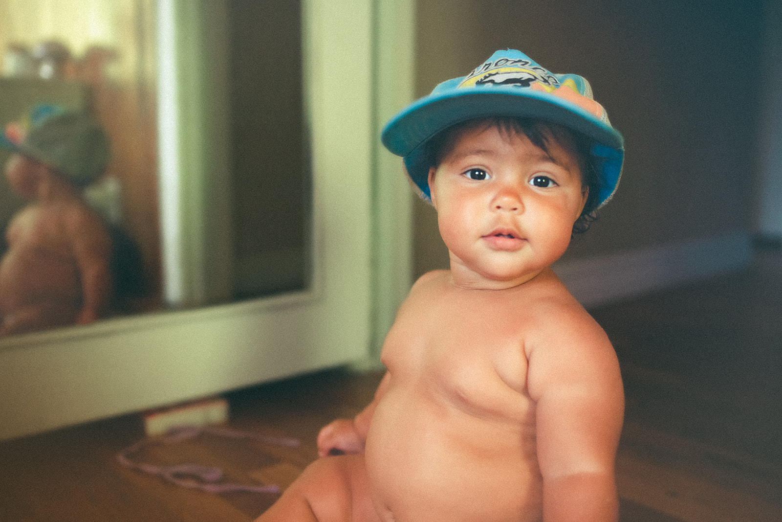 A baby sits shirtless wearing only a blue hat on the floor