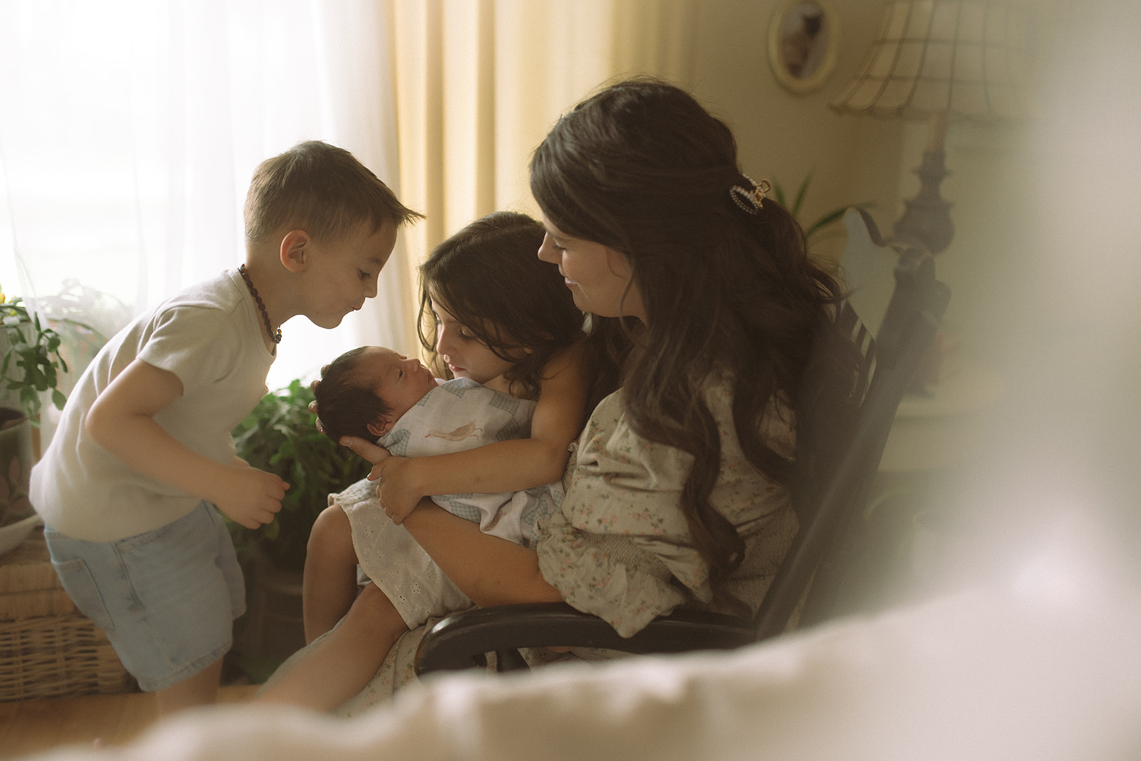 A happy mom in a floral dress sits in a rocking chair sharing her newborn with young son and daugher