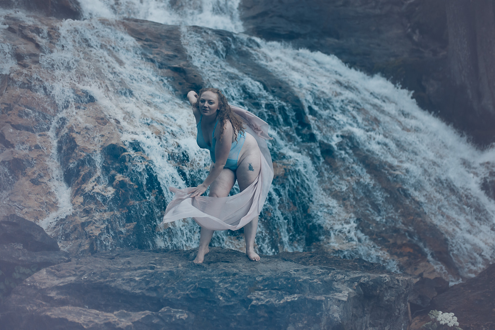 A woman with red hair in blue lingerie plays with a pink shawl in a forest by a waterfall after visiting Corvallis Spas