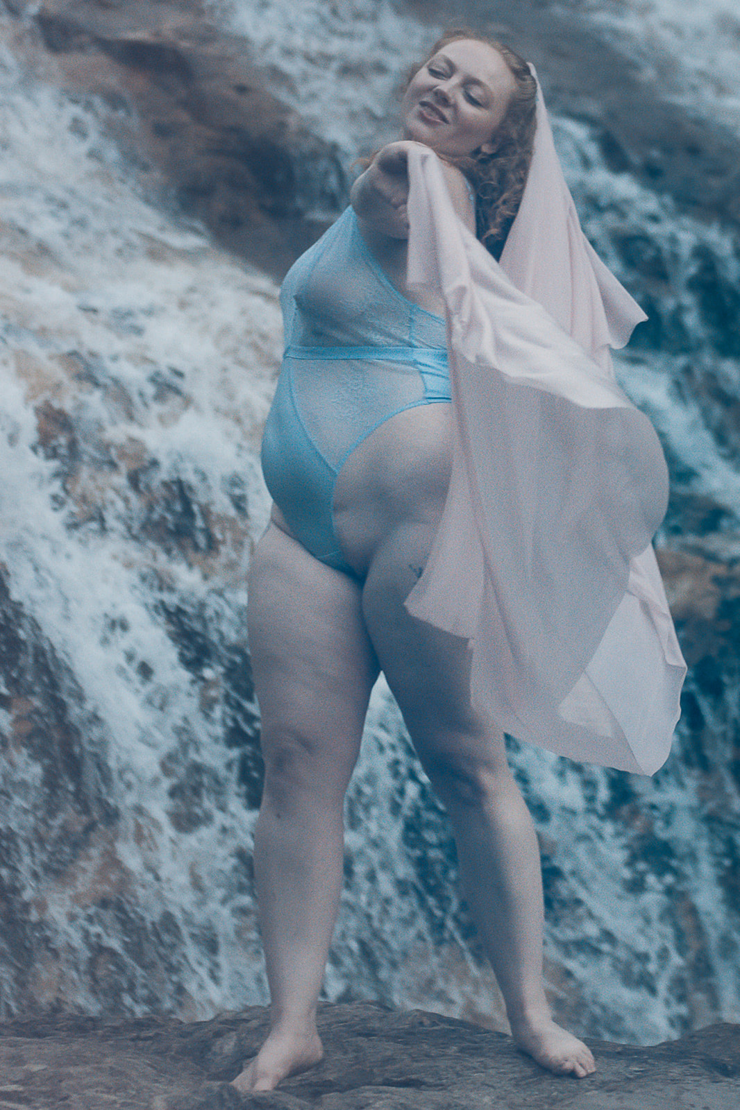 A woman in blue lingerie dances with a pink shawl on a boulder by a roaring waterfall after visiting Corvallis Spas