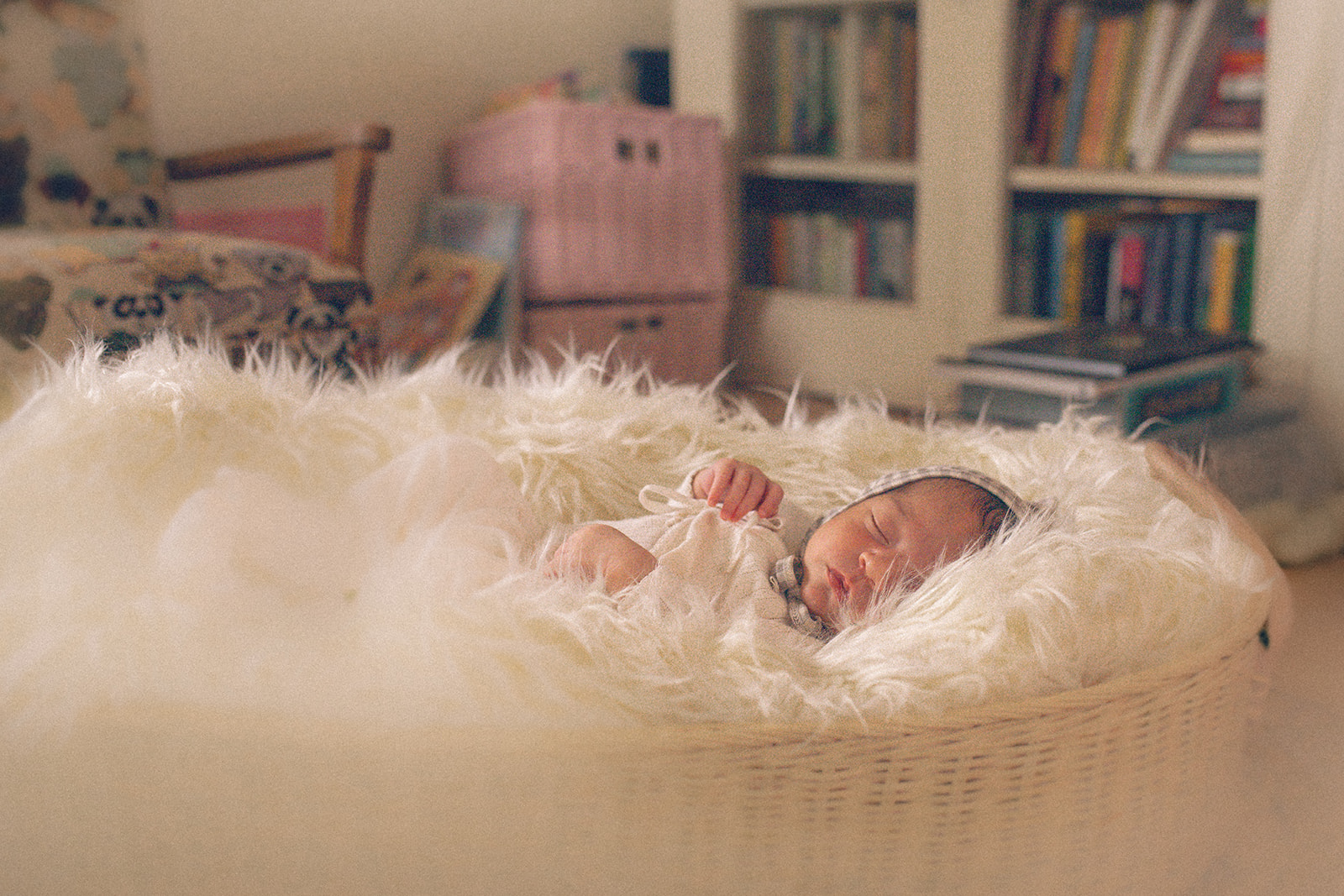 A newborn baby sleeps in a fur blanket in a wicker crib in a nursery filled with books