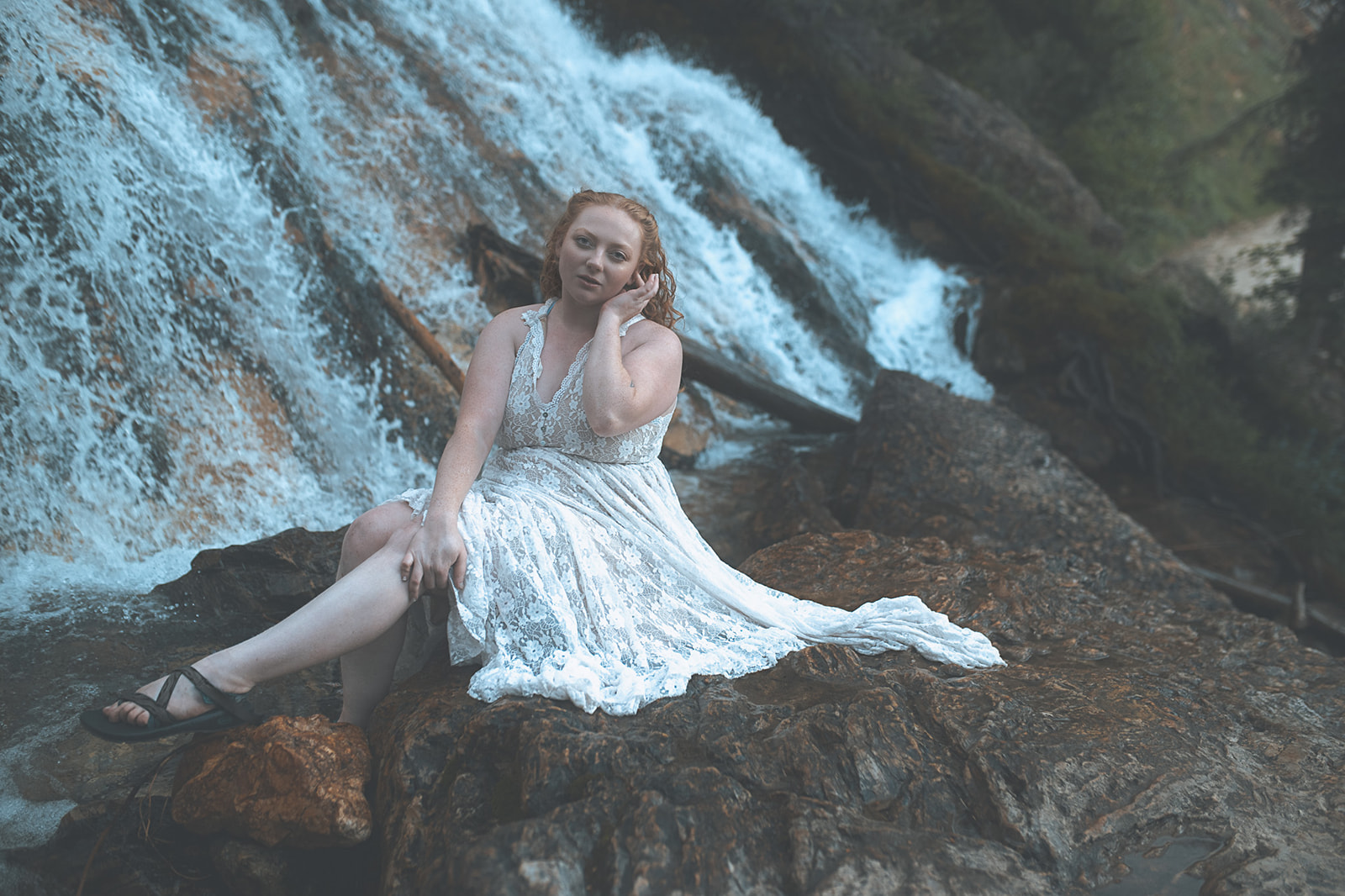 A woman with red hair and a white lace dress sits on the edge of a waterfall after visiting hair salons in Corvallis