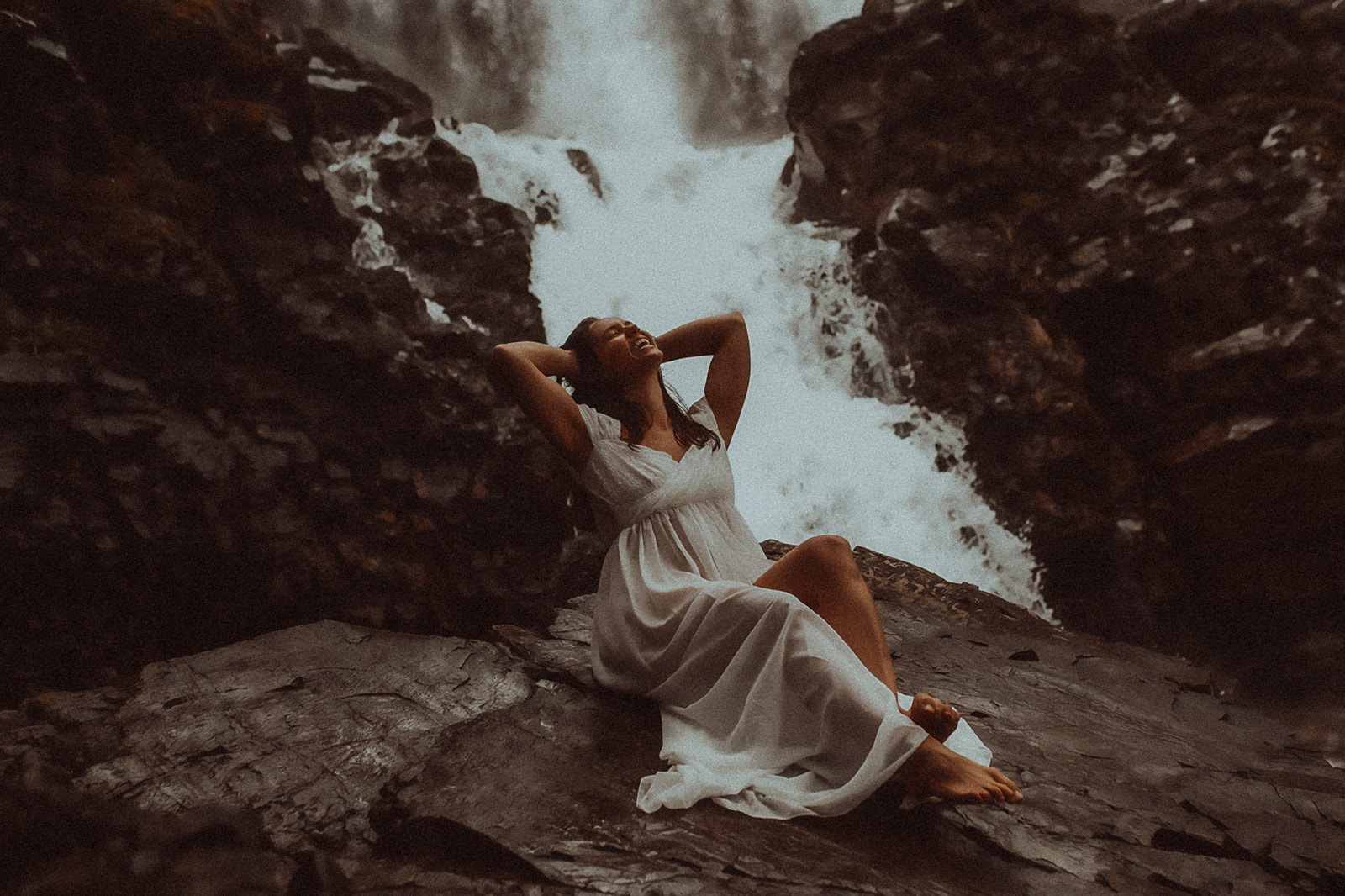 A woman leans back on a rock under a waterfall in a white gown after visiting hair salons in Montana