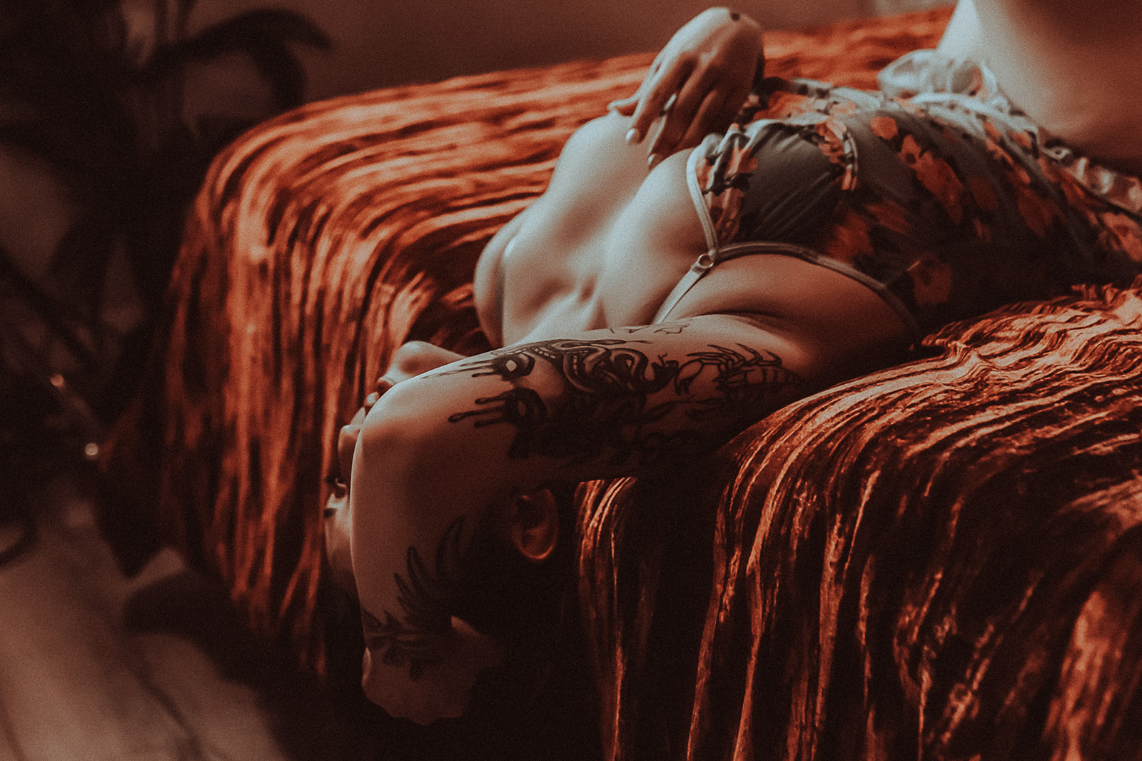 A tattoo covered woman lays back over the edge of a red bed in floral lingerie with a hand in her hair