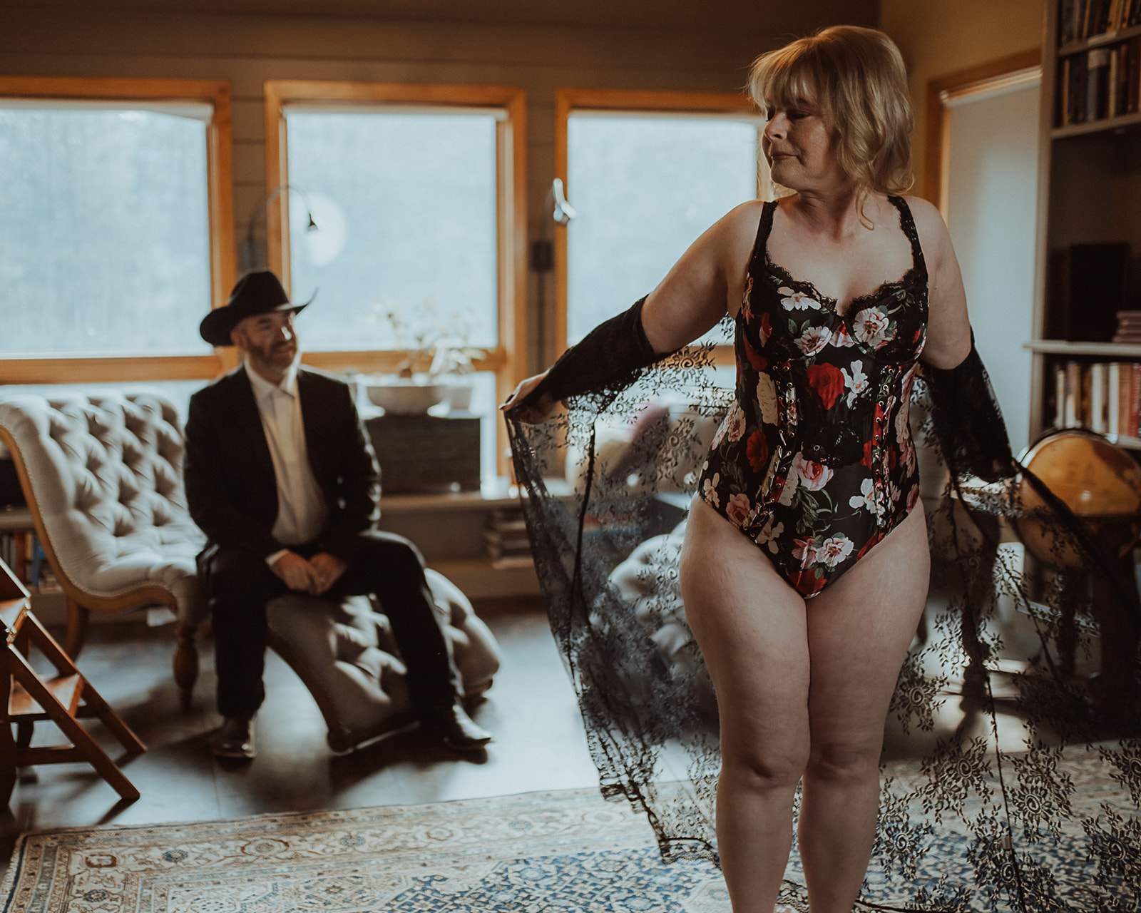 A woman in rose lingerie stands in front of her husband sitting on a lounge chair behind her after staying at Missoula Luxury Hotels