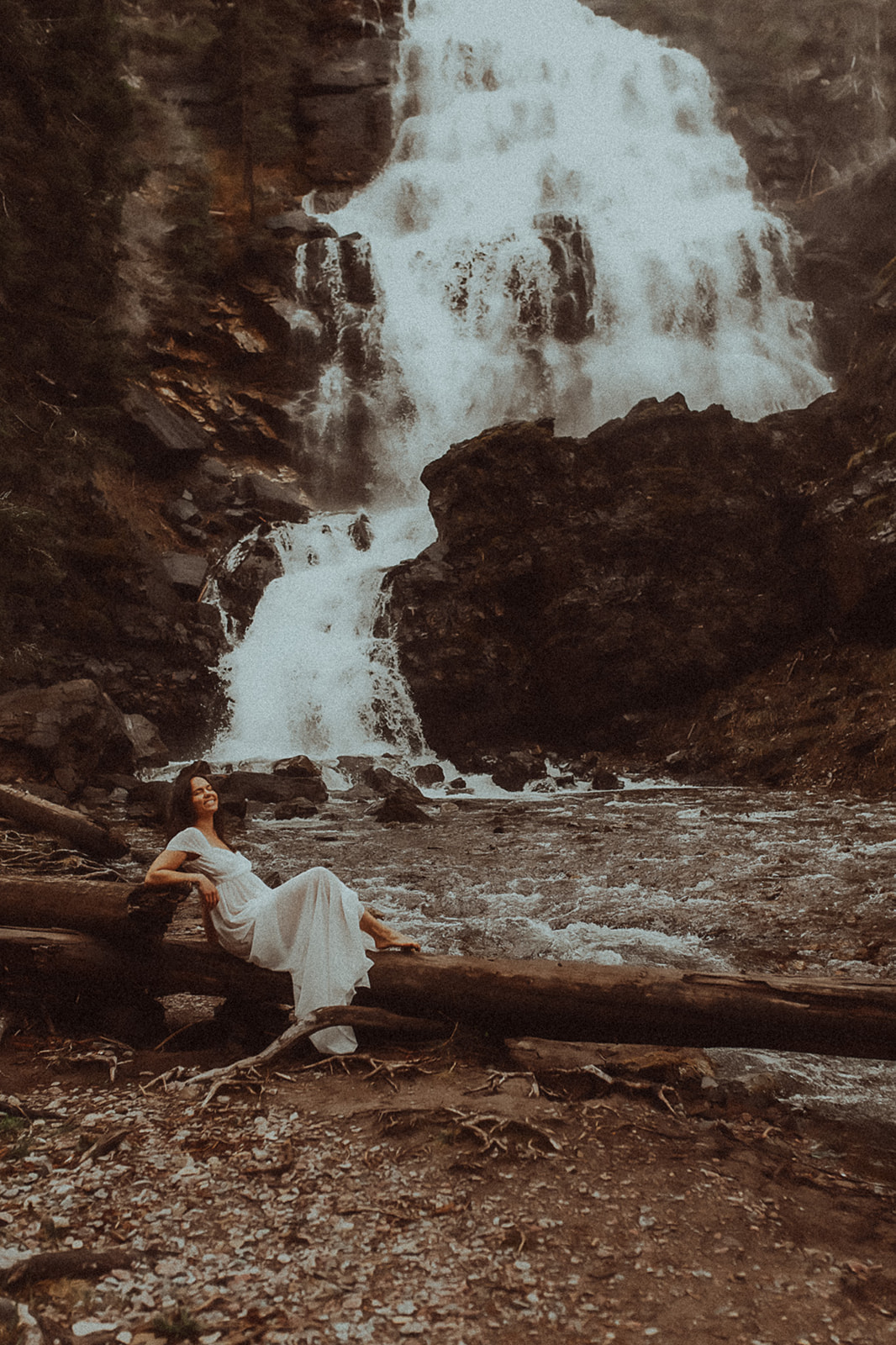 A happy brunette woman in a white dress sits on a fallen log by a large waterfall