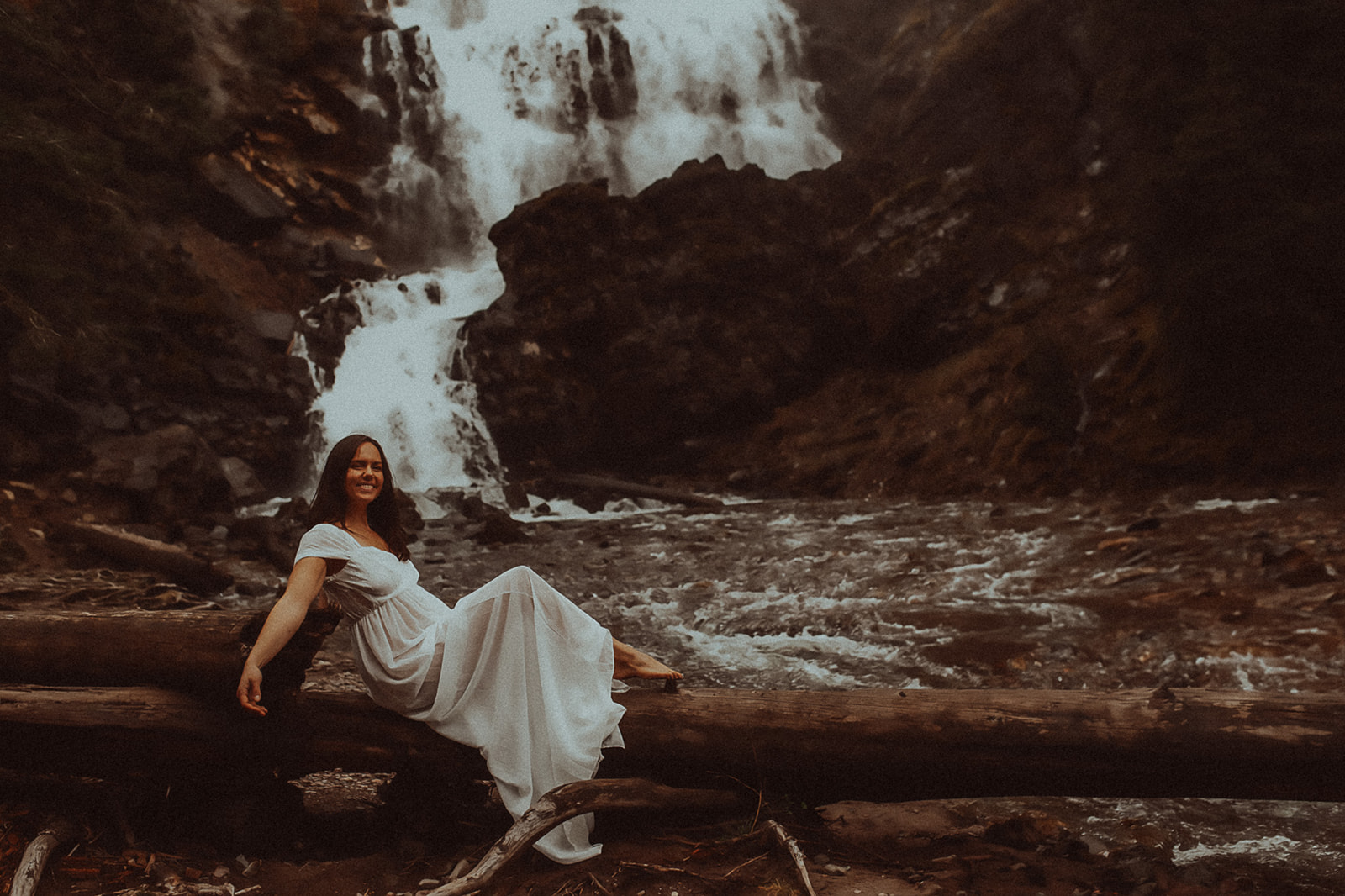 A smiling woman in a white dress sits barefoot on a fallen tree under a waterfall after visiting Missoula Spas