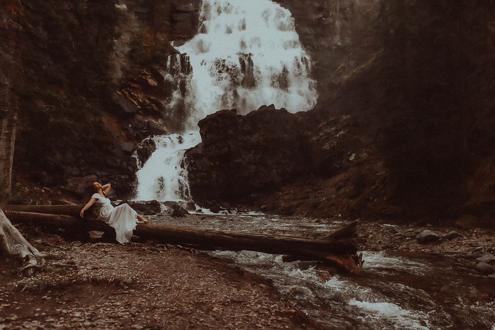 A woman in a white dress sits on a fallen tree under a waterfall after visiting Missoula Spas