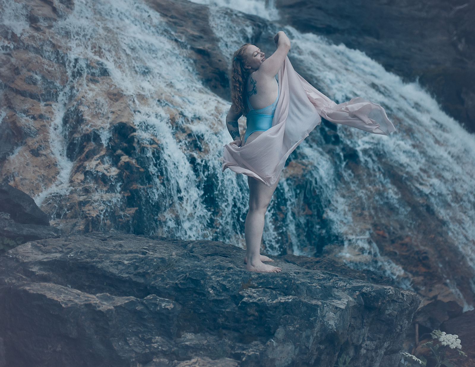 A woman dances on a boulder by a waterfall in blue lingerie with a pink coverup after some Missoula Yoga