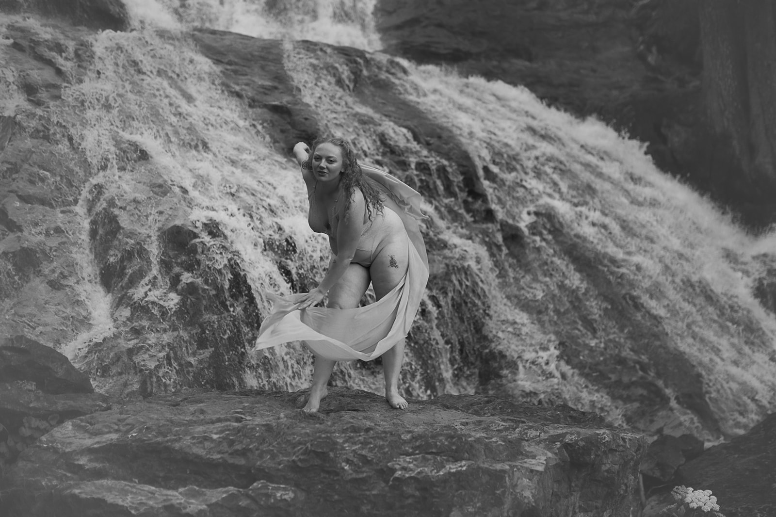 A woman in lingerie dances with some fabric on a rock on a waterfall