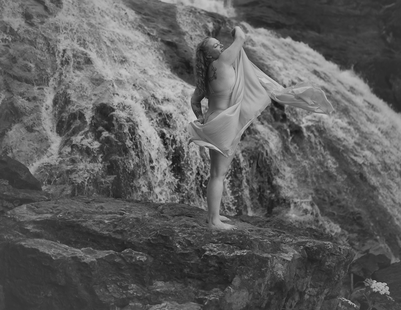 A woman dances on a rock under a waterfall in her underwear with a shear fabric after some Montana Yoga
