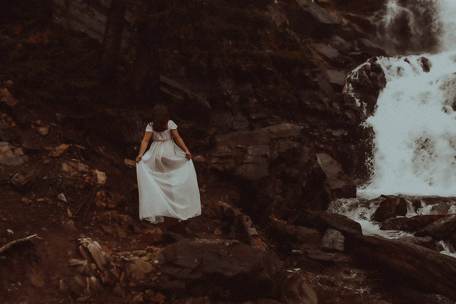A woman holds her white dress while walking to the edge of a waterfall