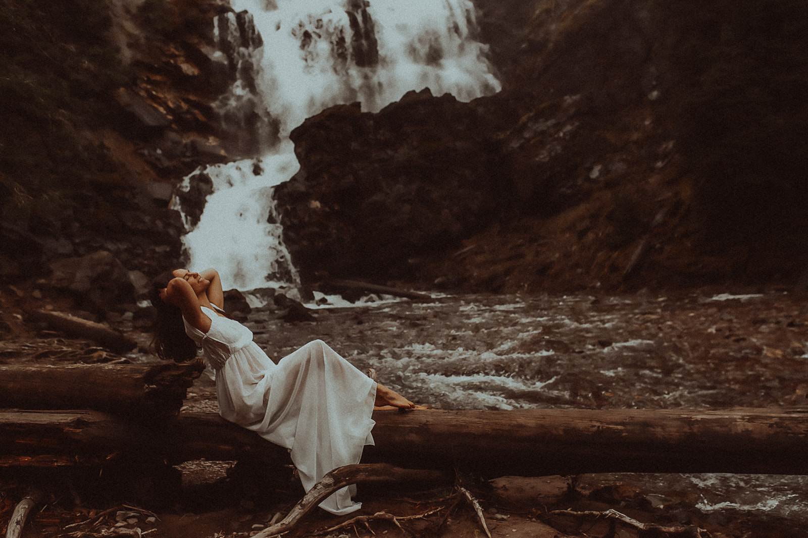 A woman in a white gown lounges on a fallen tree over a river by a waterfall after visiting nail salons in Missoula, MT