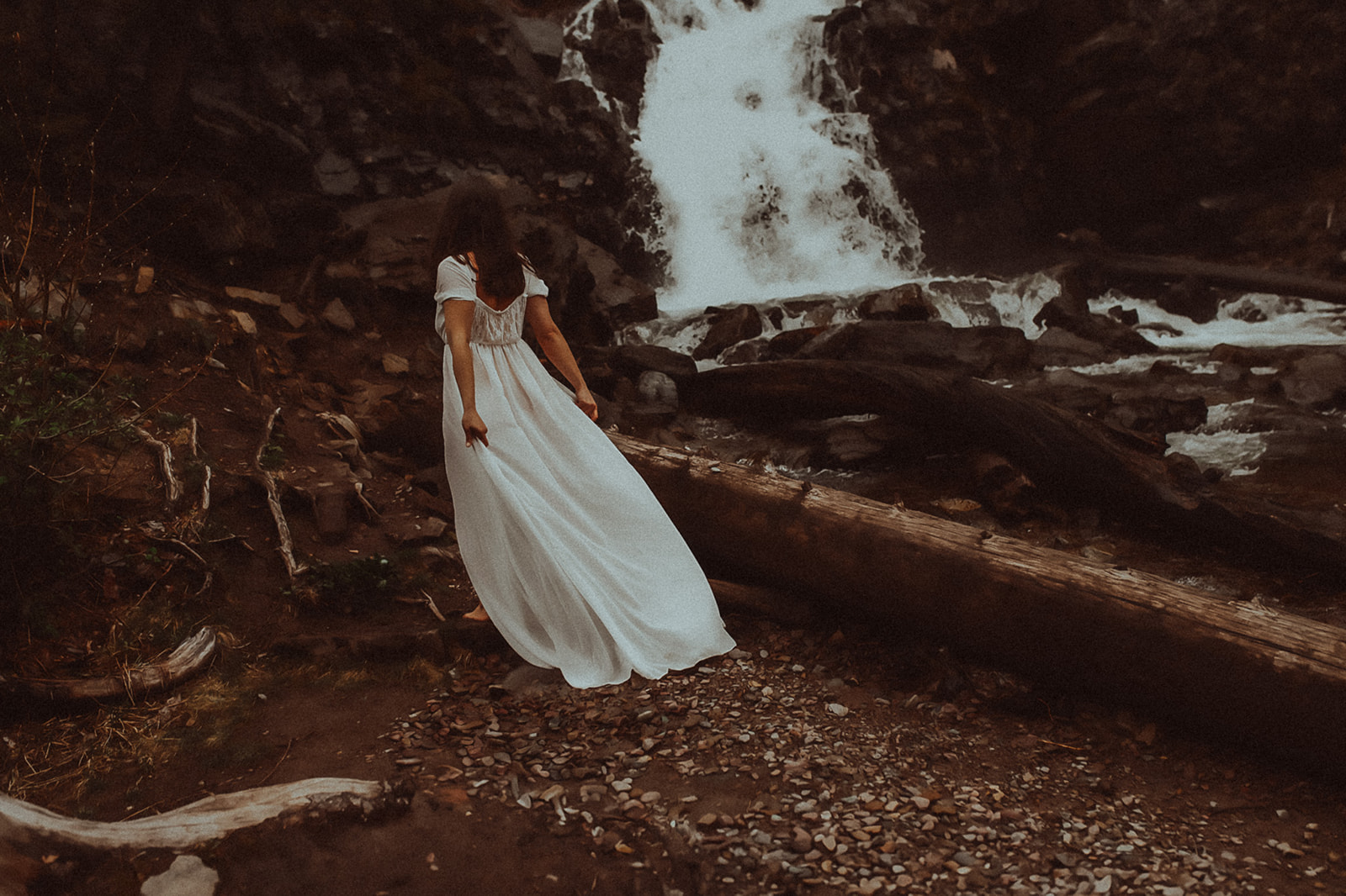 A woman in a white gown walks on the edge of a river by a waterfall after visiting nail salons in Missoula, MT