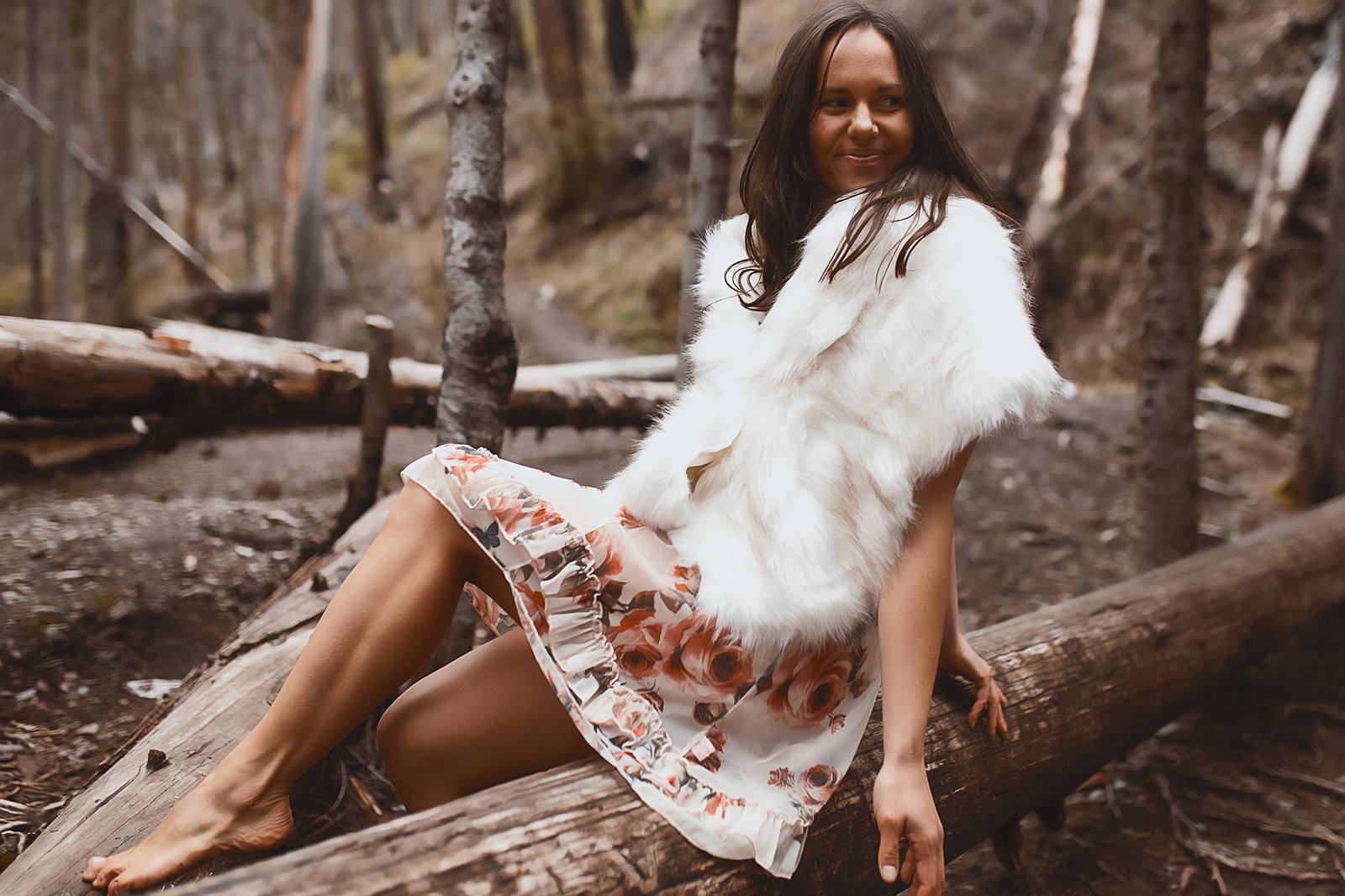 A woman in a floral gown and fur cover sits on a log in a forest smiling after visiting tanning salons in Missoula, MT