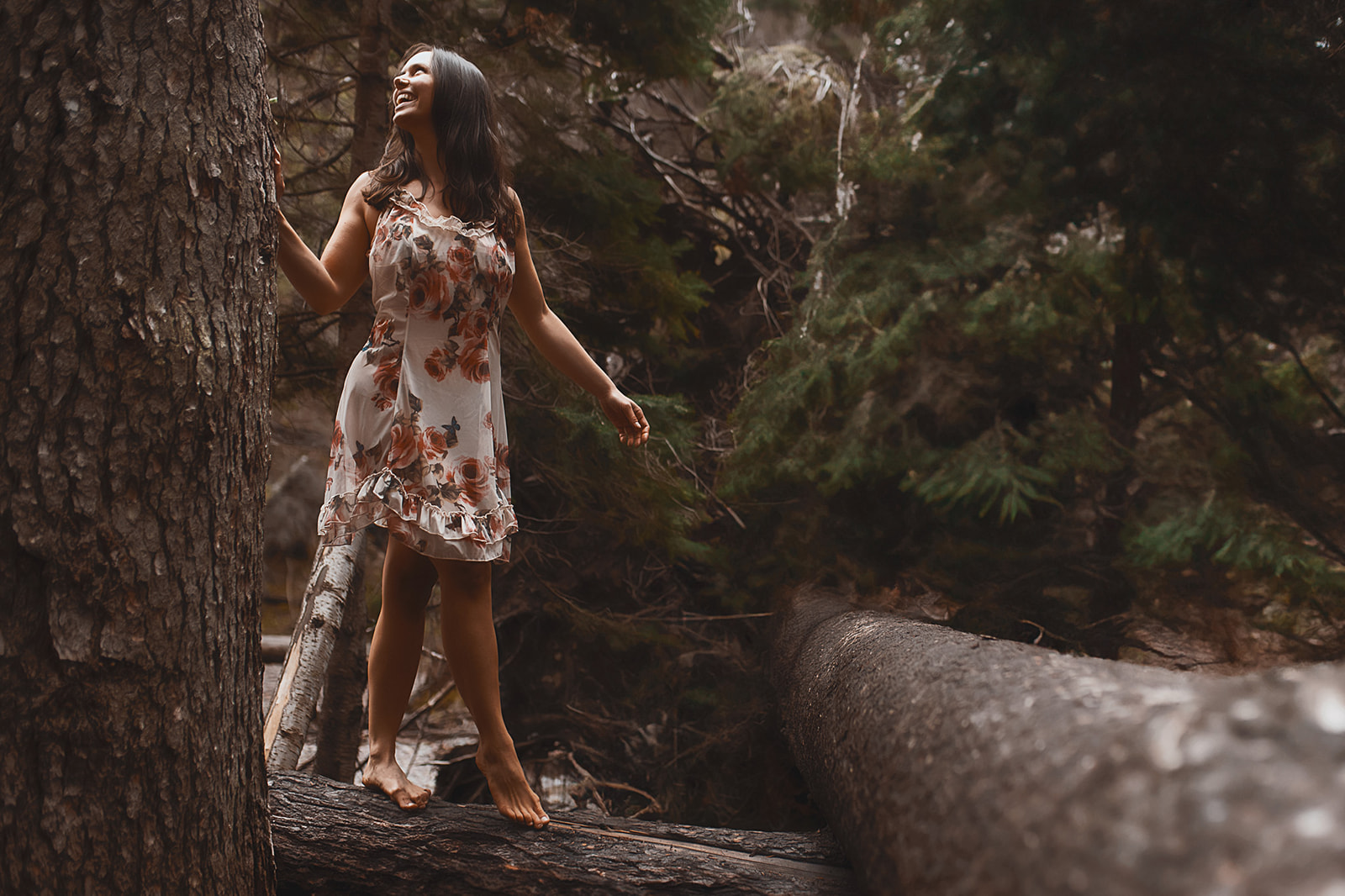 A woman in a floral gown walks on a fallen tree in a forest after finding tanning salons in Missoula, MT
